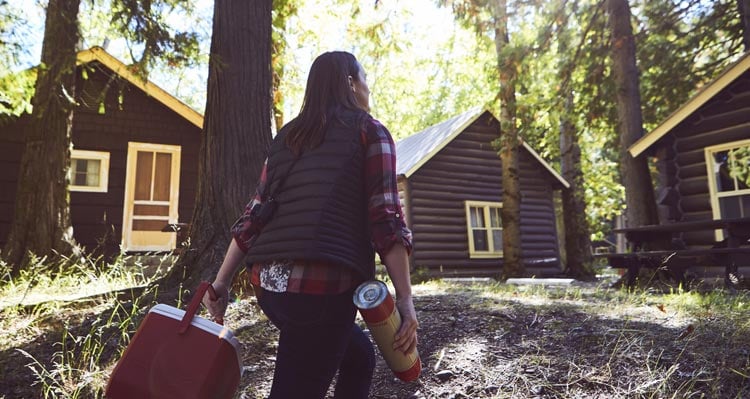 A woman carrying a thermos and cooler walks towards cabins set among trees