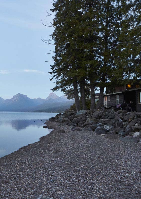 A rustic cabin sits at the shore of a serene lake, with mountains across.