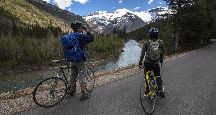 Two cyclists stop to enjoy and take photos of a view of a creek and snow-covered mountains.