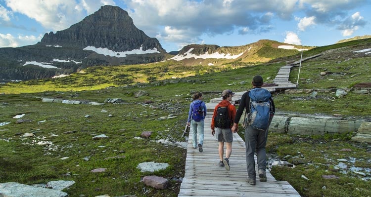 Three people walk along a boardwalk in an green, mountainous meadow