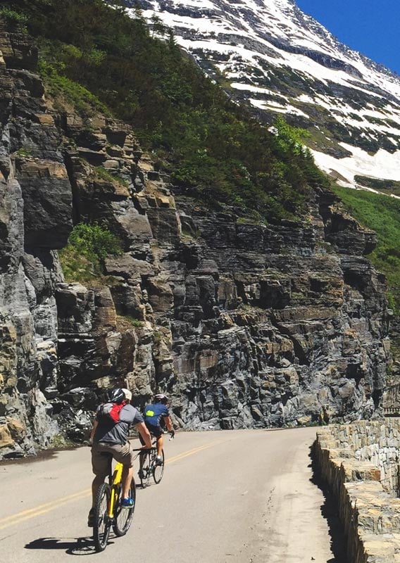 Two cyclists ride on a mountain road towards a waterfall.