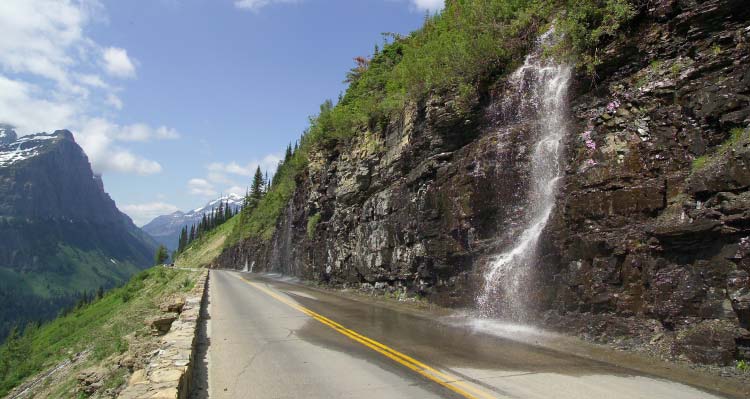 A waterfall splashes into a road running alongside a mountain.