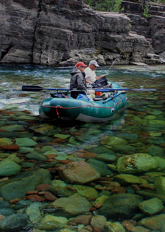 Two men float in a fishing boat along a shallow river filled with green and red rocks.