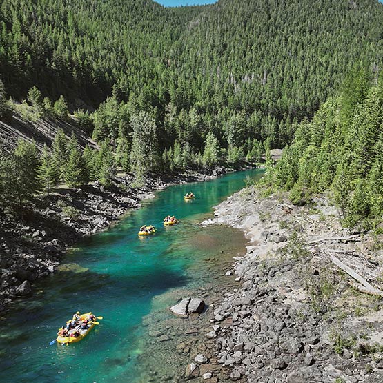 A calm river with trees and mountains on either side. Four rafts float down.