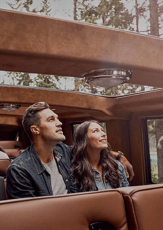 Two people in a historic style touring automobile, looking up out a glass roof.