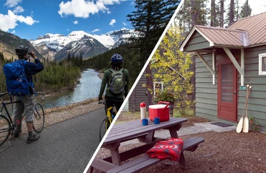 Two cyclists stopped at the roadside next to a wide river below mountains. A green cabin and picnic.