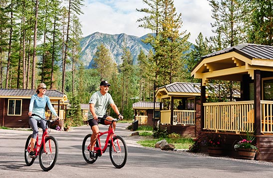 Two people ride bikes between brown cabins under tall conifer trees.