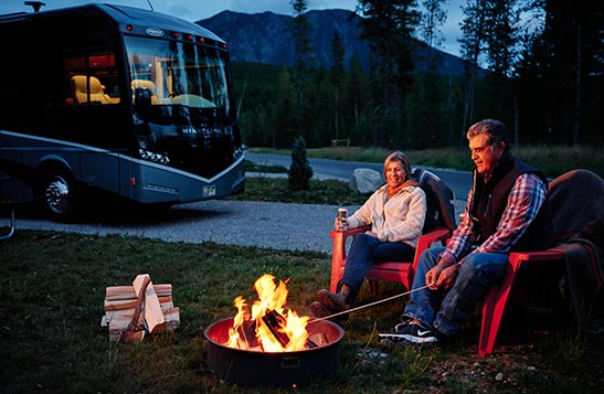An older couple are sitting outside next to a firepit with their RV parked close at night.