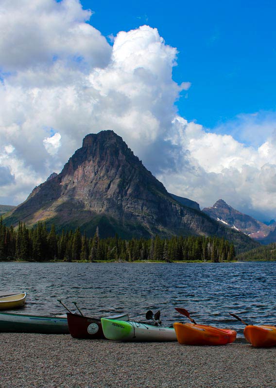 A row of canoes and kayaks at the shore of a blue lake below tree-covered mountains.