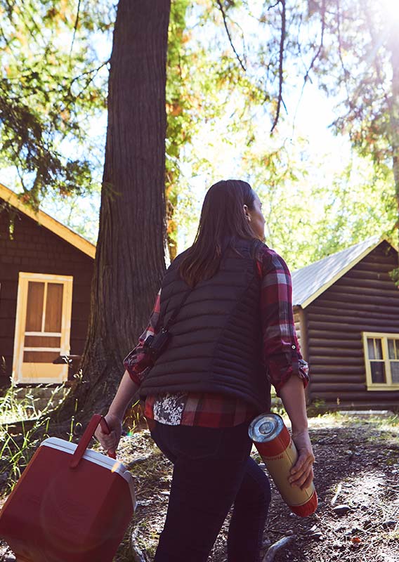 A woman walks towards a series of wooden cabins nested between tall trees.