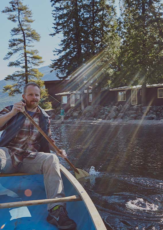 A man canoes away from a lakeshore with small cabins under conifer trees.