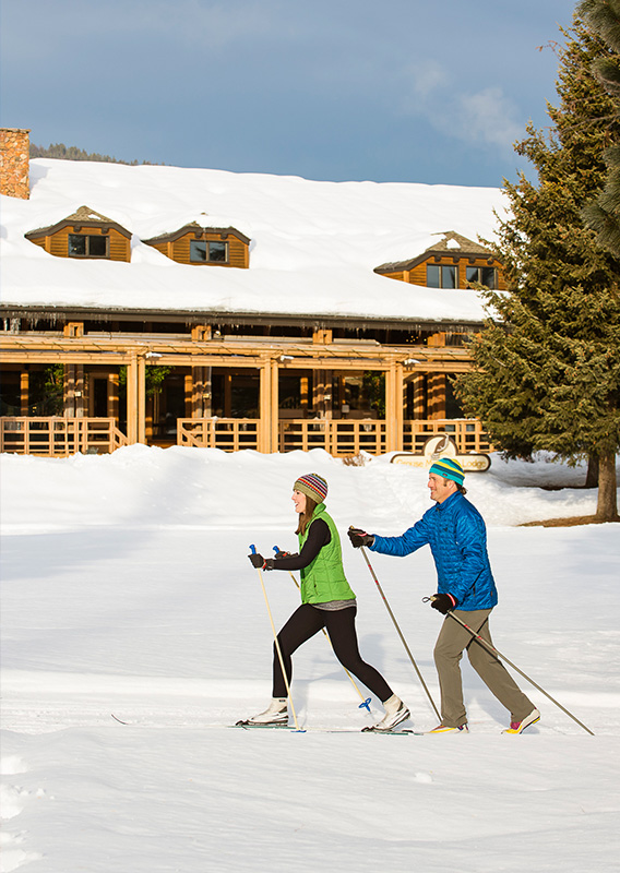 Two cross-country skiiers ski past a large lodge on a sunny winter day
