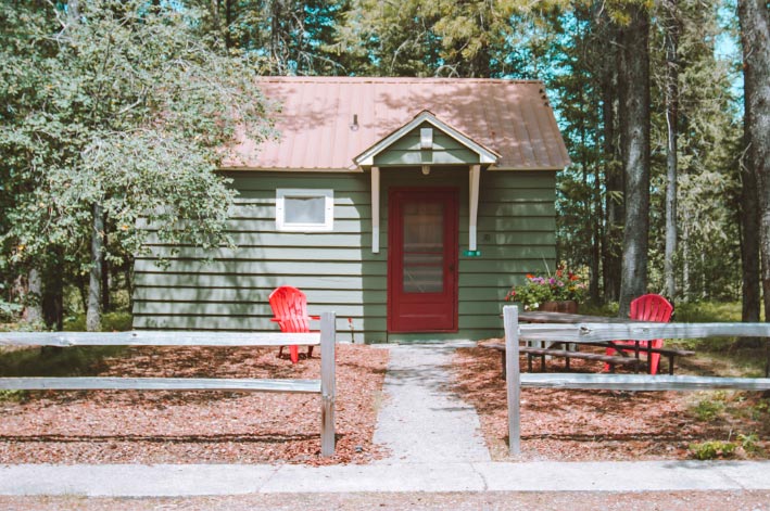 A green wooden cabin and two red Adirondack chairs shaded by trees, behind a small wooden fence.