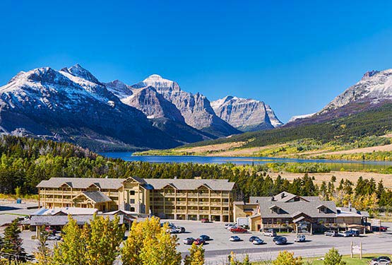 St. Mary Village sits against a forest, ahead of a blue lake and high mountains.