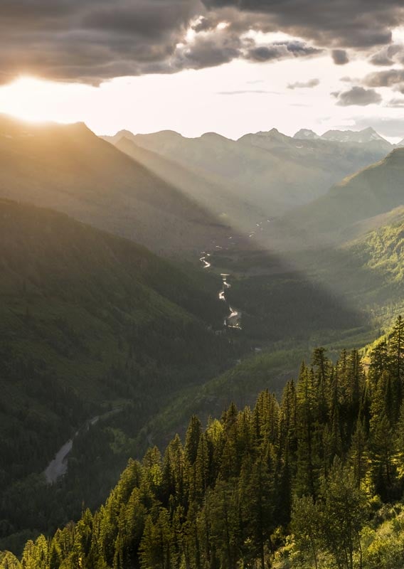 The sun sets behind mountains, shining into a wide, green valley