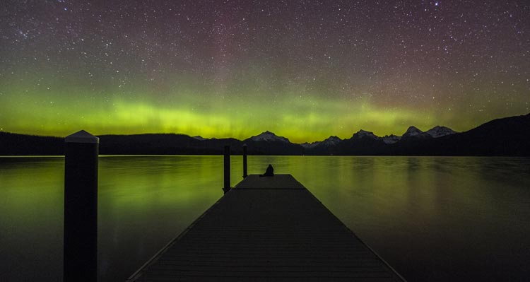 A view down a dock looking towards mountains and green aurora.