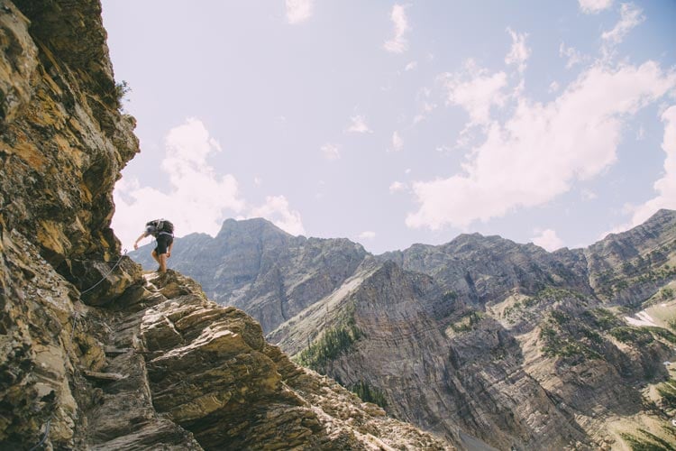 A person holds onto a metal cable climbing high above mountains