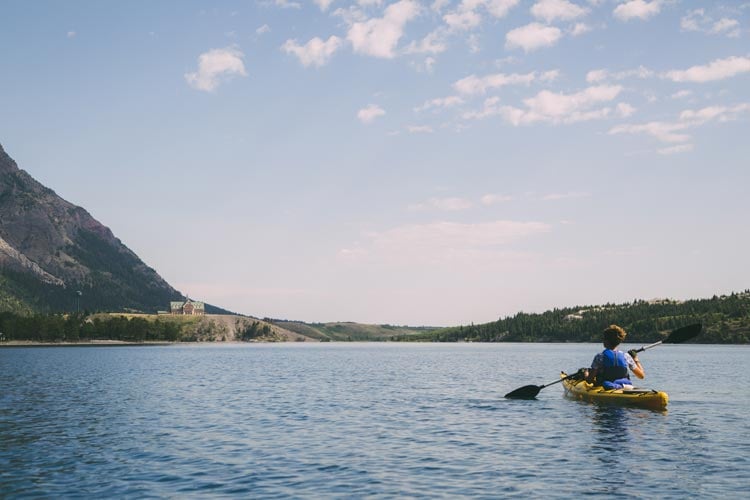 A kayaker paddles on a lake towards the Prince of Wales Hotel, at the meeting of grasslands and mountains