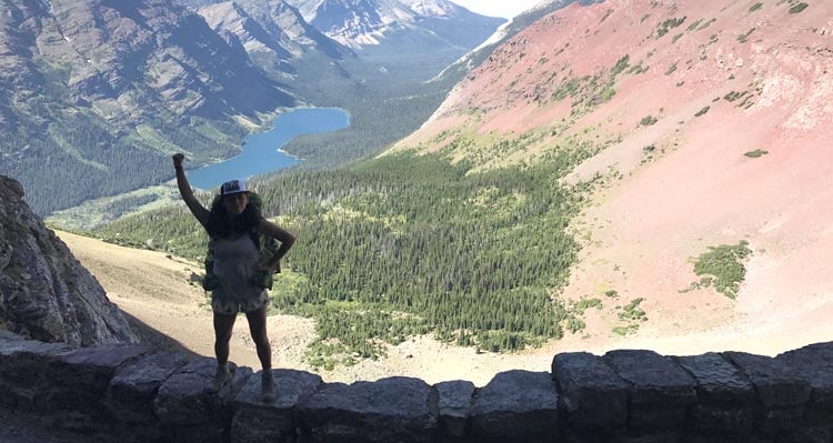 A hiker raises a hand at a view point high above a blue mountain lake and a wide valley.