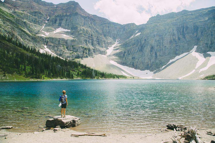 A person stands on a rock at the edge of a blue lake surrounded by high mountain cliffs