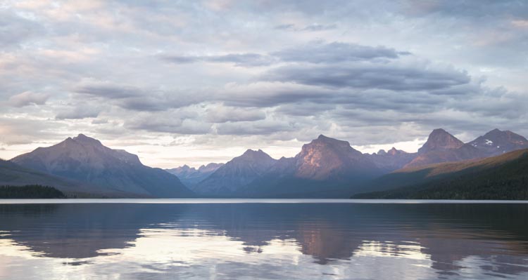 The setting sun shines on rocky and tree-covered mountain faces across a calm, clear lake.