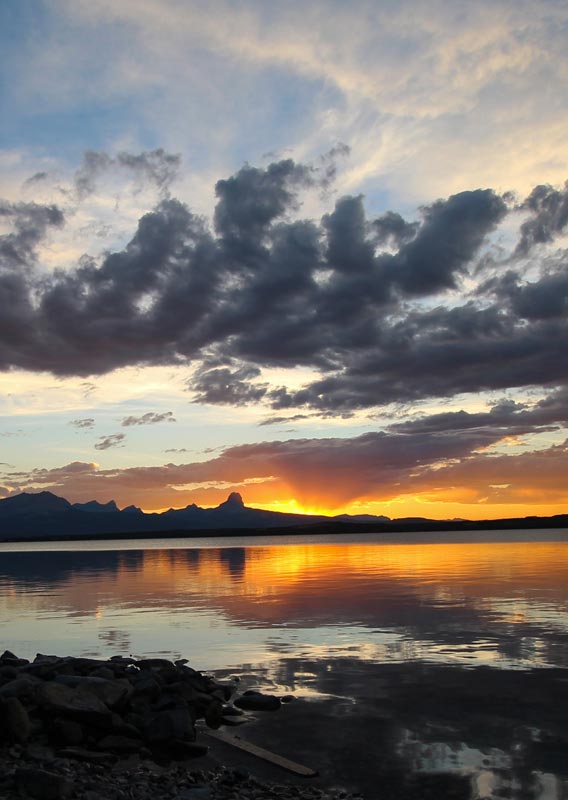 The orange glow of the setting sun shines between mountains and plains behind a calm lake.