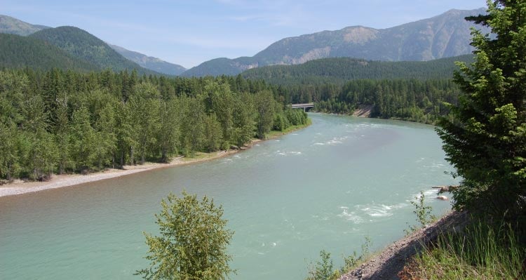 A person fly-fishes in a river that flows between tree-covered hills.