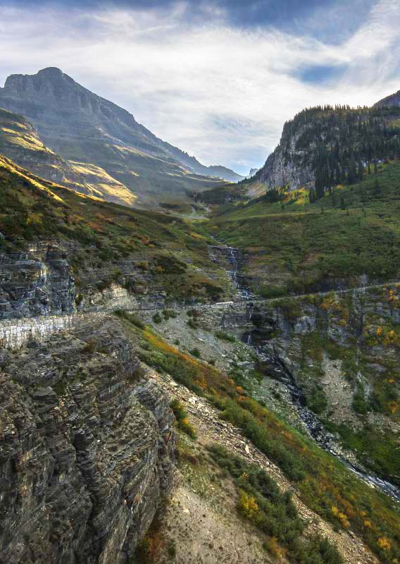 A streaming waterfall cascades down an alpine mountainside towards and beneath a road.