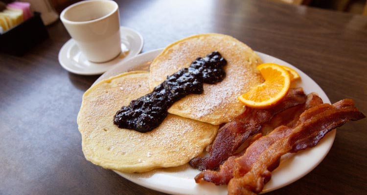 A breakfast of pancakes and jam with bacon an orange slice and coffee cup.