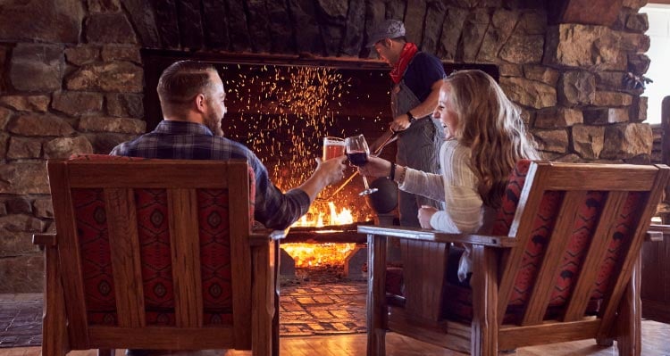 A couple cheers a drink next to a roaring fireplace.