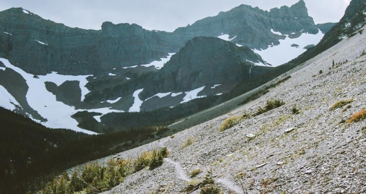 A trail traverses a scree slope, and looking towards a snow-covered mountainside.