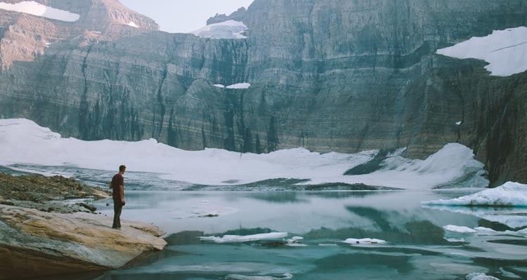 A hiker stands at the edge of a mountain lake surrounded by rocky cliffs and ice.