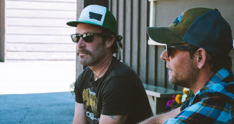 Two men in baseball caps sit on a porch.