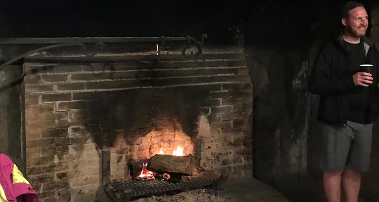 A man stands next to a rustic fireplace.