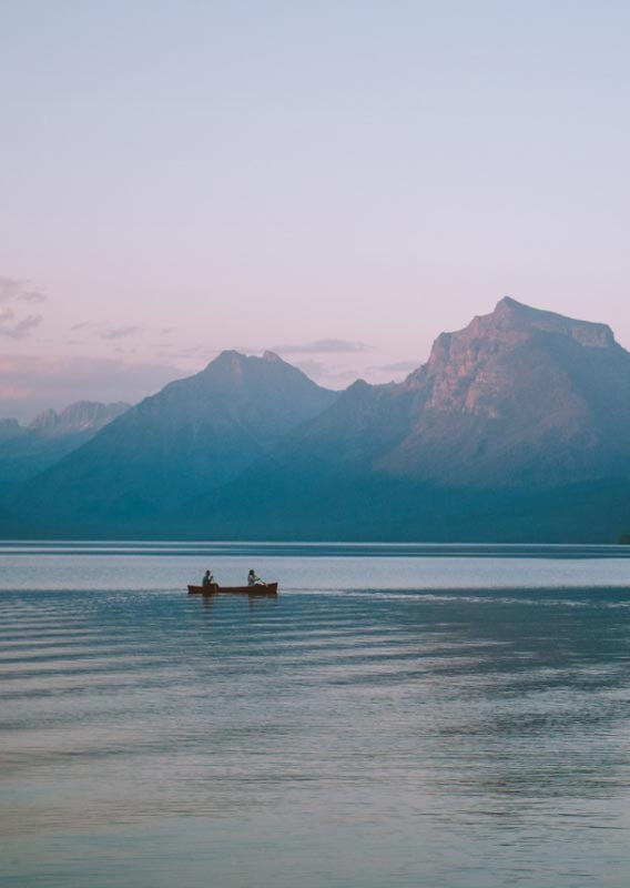 Two canoers paddle on a clear lake with mountains glowing in a sunset.