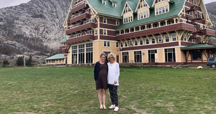 Two women stand close together in front of the Prince of Wales Hotel, near a mountainside.