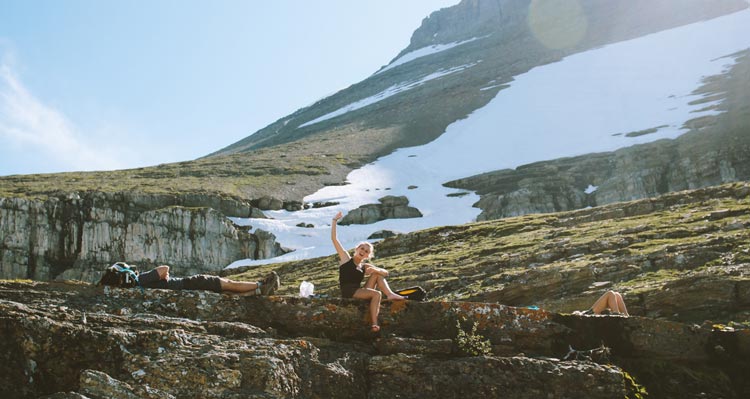 A hiker waves from a rocky ledge in front of a snow-covered mountainside.