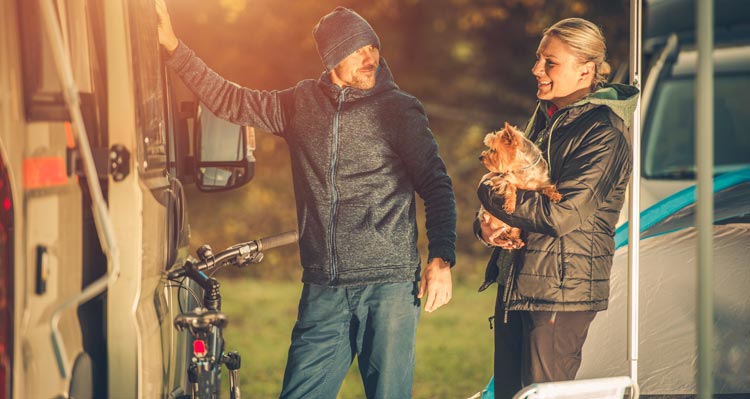 Two people stand outside their RV with a small dog.