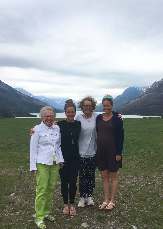 Four women embrace and pose for a photo on a grassy plain above a lake.