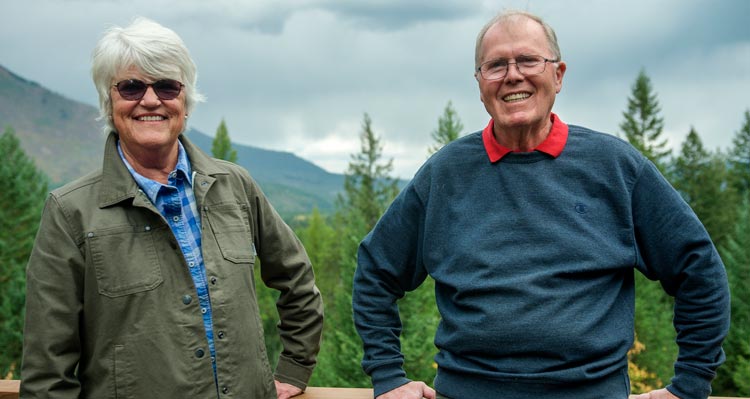 Two people stand at a deck in front of a forest and mountainside