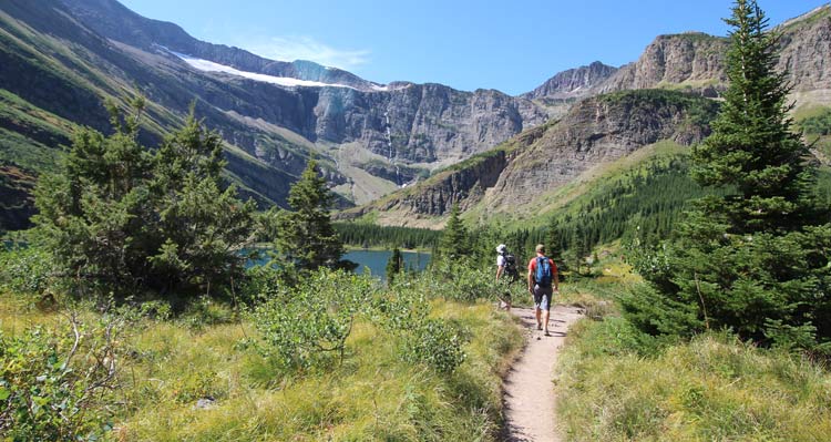 Two hikers walk towards a lake surrounded by rocky cliffs.