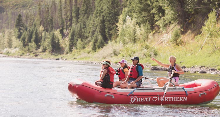 A group of people sit on a raft on a wide river with tree-covered banks.