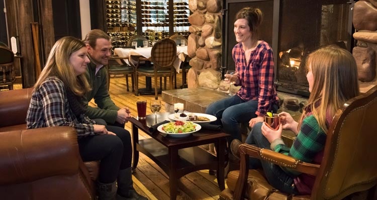 Four people sit around a table by a fireplace.