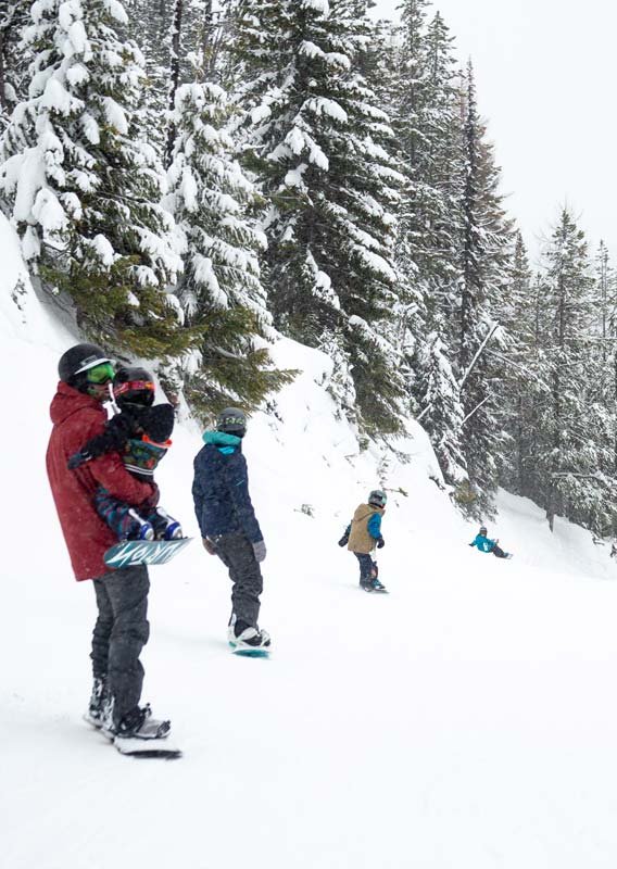 A family snowboards along a gentle slope alongside a snowy forest