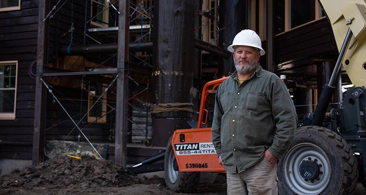 A man stands in construction clothing and gear outside a large wooden building.