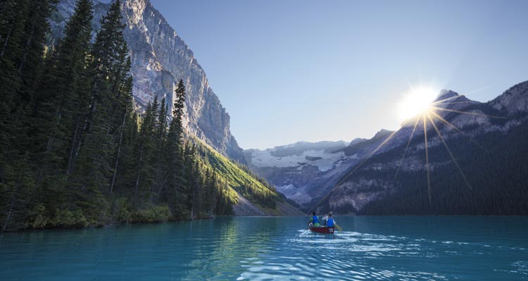 A pair of canoers paddle on a blue lake surrounded by forest-covered mountains.