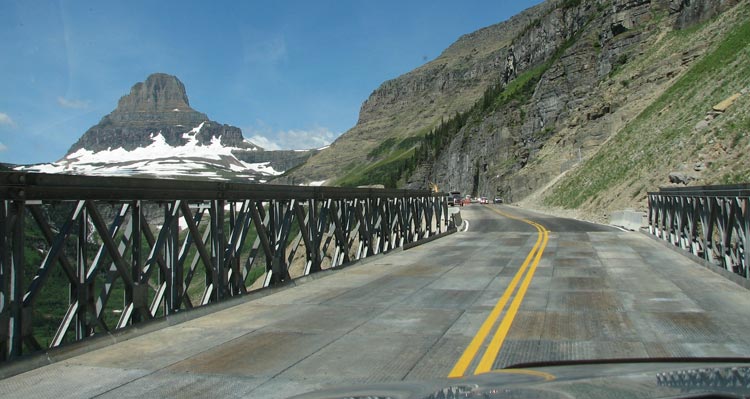 A view out the front window of a car on the Going-to-the-Sun Road looking towards mountains.