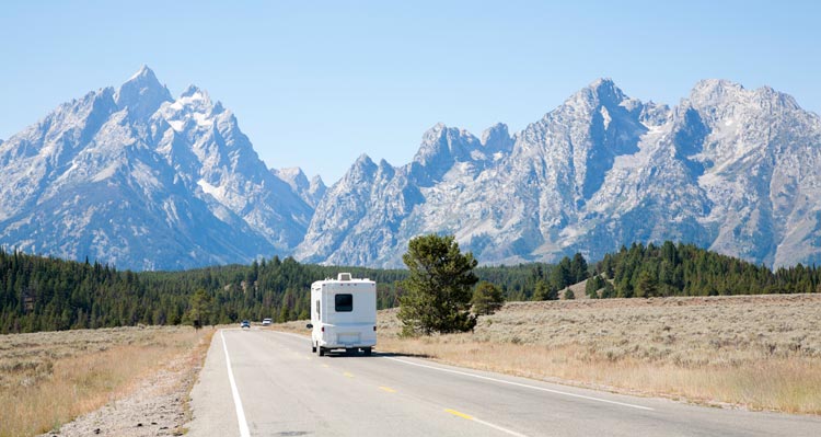 An RV drives on an open road towards jagged mountain peaks.