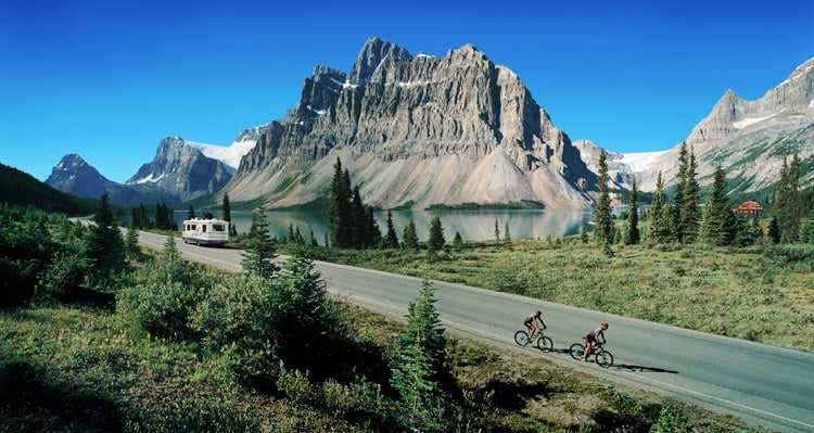 An RV drives on a road alongside a lake and tall mountains.