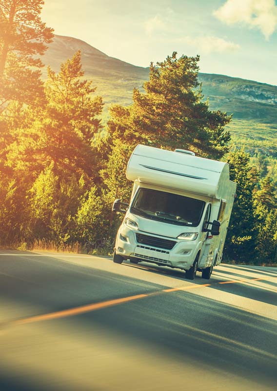 An RV drives down a sunny road below forested hillsides.